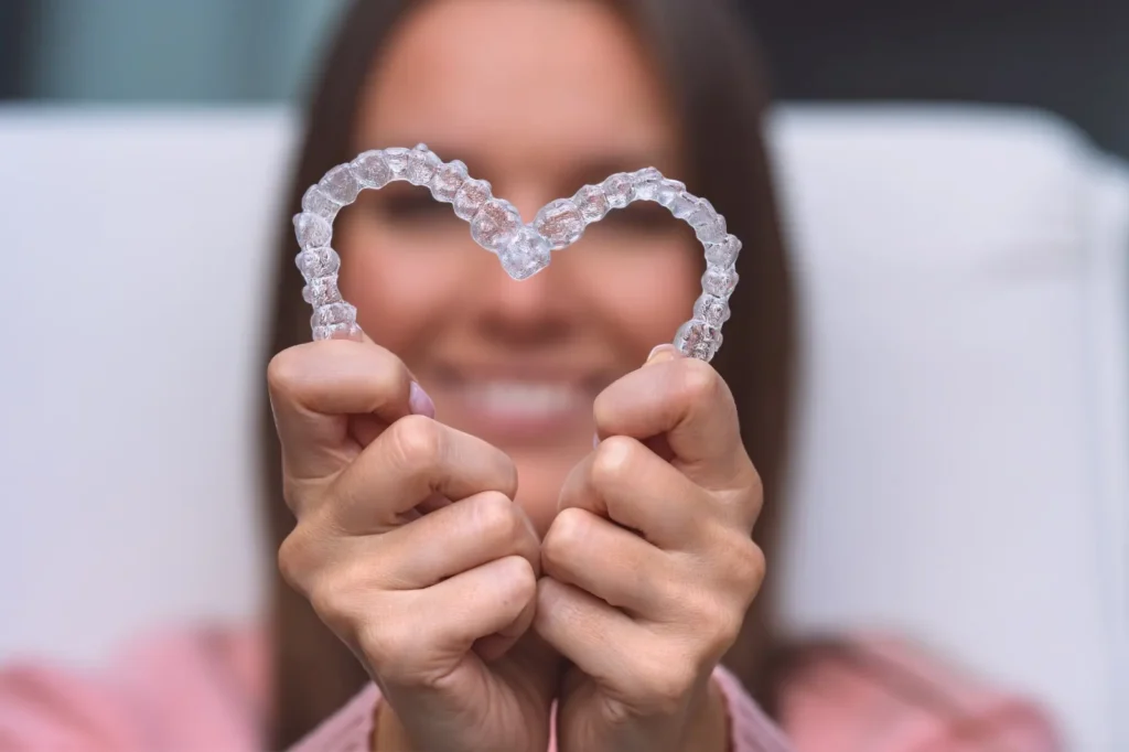 Woman holding transparent Invisalign aligners shaped like a heart while smiling.